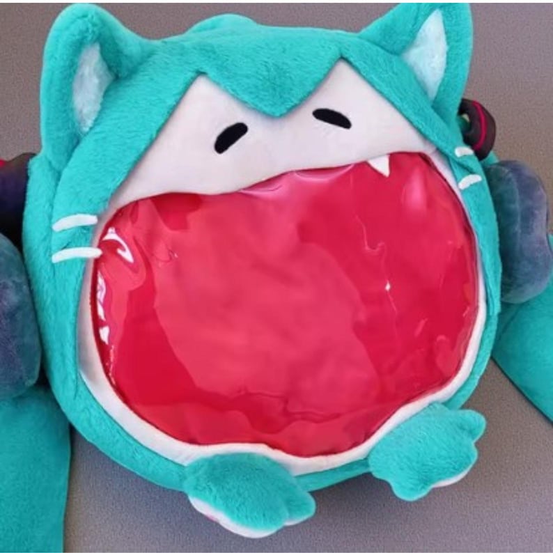 May include: A teal plush backpack shaped like a cartoon cat's head, featuring white accents, black eyes, and a large, open red mouth. The backpack has cat ears and paws, with a clear, plastic-like opening for storage.