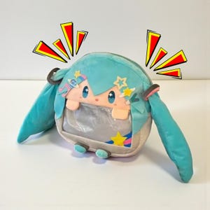May include: A plush, round, light blue character bag with long pigtails. The bag has a clear window showing a face with large blue eyes and star accents. The bag is decorated with star and bow details.