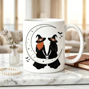 Puede incluir: Una taza de cerámica blanca con un gráfico negro de dos figuras con sombreros de bruja sentadas en una luna creciente, tomadas de la mano. Las figuras tienen cabello naranja y negro, con estrellas y murciélagos en tinta negra.