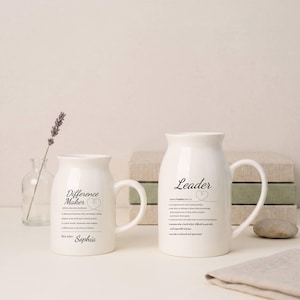 Puede incluir: Dos tazas de cerámica blanca con texto negro. Una taza dice "Difference Maker" y la otra dice "Leader". Ambas tazas tienen un asa curva y una base ancha. Un pequeño jarrón de cristal con una ramita de lavanda está en el fondo.