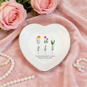 Puede incluir: Plato de cerámica blanco en forma de corazón con borde dorado. El plato presenta ilustraciones florales y los nombres Alexis, Evelyn y Stella. El texto dice: "Las hermanas son flores diferentes del mismo jardín".