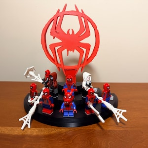 Op de afbeelding: Een display met verschillende Spider-Man Lego-figuren, waaronder Miles Morales en Spider-Gwen, gerangschikt op een zwarte cirkelvormige basis. Een groot rood Spider-Man-logo is achter de figuren geplaatst. De figuren staan in verschillende poses.
