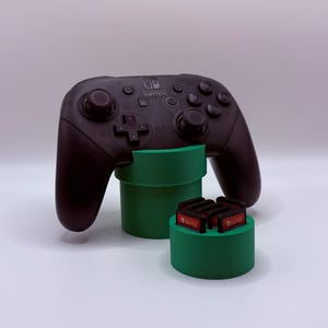 May include: A black Nintendo Switch Pro controller resting on a green cylindrical stand, accompanied by a smaller green cylindrical holder containing several Nintendo Switch game cartridges with visible red and white labels.