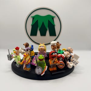 May include: A circular black display stand featuring a collection of Muppet-themed minifigures, including Kermit the Frog with a banjo, Miss Piggy, and Animal with drums. A white circular sign with a green letter M logo stands in the center.