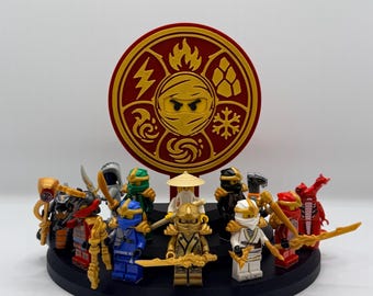 May include: A circular black display stand holding eleven plastic ninja minifigures in various colors including blue, gold, white, and red. Behind the figures stands a large red and gold circular emblem featuring elemental symbols and a central ninja mask.