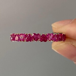Könnte beinhalten: Ein goldener Ring mit einer Reihe herzförmiger, leuchtend rosa Edelsteine. Die Edelsteine sind eng aneinander gesetzt und bilden eine durchgehende Reihe von Herzen. Der Ring wird vor einem neutralen grauen Hintergrund präsentiert.