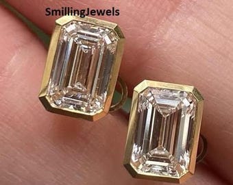 May include: A pair of gold stud earrings featuring emerald-cut, clear gemstones. The earrings have a rectangular shape with a gold border. The earrings are displayed on a light-coloured background.