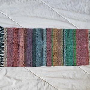 May include: A rectangular, handwoven textile with vertical stripes in various colors, including red, purple, green, and blue. The ends have a fringe of dark green yarn. The textile is likely a small rug or decorative piece.