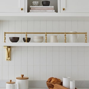 Op de afbeelding: Een keukenkast met diverse items. Boven de plank is een messingkleurige metalen stang gemonteerd, met een verzameling kommen, mokken en een kan. De plank toont ook witte kommen, een witte mok en een witte kan. De achtergrond is een witte betegelde muur.