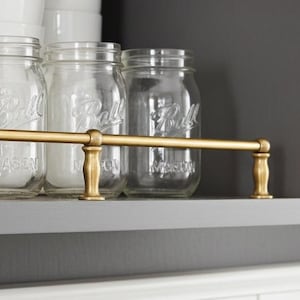 May include: A cabinet shelf displays clear glass Ball mason jars, white bowls, and a jar with a wooden lid. A gold-colored metal bar runs across the front of the shelf. The cabinet is painted a dark gray color.
