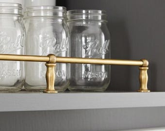 May include: A cabinet shelf displays clear glass Ball mason jars, white bowls, and a jar with a wooden lid. A gold-colored metal bar runs across the front of the shelf. The cabinet is painted a dark gray color.