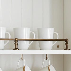 Peut inclure: Une armoire de cuisine blanche avec une barre de couleur laiton contenant six tasses blanches. En dessous, six tasses blanches avec des bords dorés sont suspendues à des crochets. Une pile d'assiettes blanches, un pot blanc et un petit récipient en céramique sont sur l'étagère du bas.