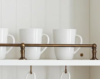 May include: A white kitchen cabinet with a brass-coloured rod holding six white mugs. Below, six white mugs with gold rims hang from hooks. A stack of white plates, a white canister, and a small ceramic container are on the bottom shelf.