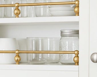 May include: White kitchen cabinet with two shelves. The shelves feature gold-colored metal rails with decorative ball accents. Various clear glass jars, white cups, and vases are displayed on the shelves.