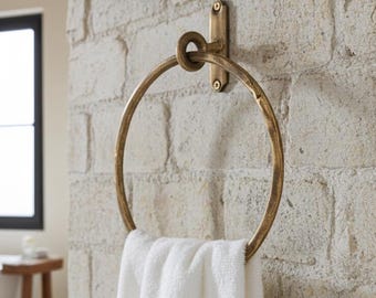 May include: A brass-colored towel ring is mounted on a textured, light-colored brick wall. A white towel hangs from the ring. The bathroom setting includes a white bathtub, a small wooden stool, and a window with a black frame.