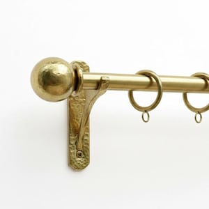 May include: A gold-coloured curtain rail with a hammered texture. The rail features a spherical finial, decorative bracket, and several hanging rings. The rail is designed to hold curtains and drapes. The hardware is made of metal.