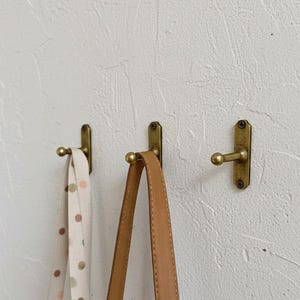 Op de afbeelding: Drie antieke messing wandhaken aan een getextureerde witte muur. Twee haken houden een lichtgekleurde band met bruine stippen en een bruinleren band vast. De haken hebben een eenvoudig, functioneel ontwerp.