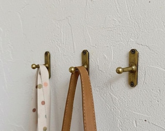 May include: Three antique brass wall hooks mounted on a textured white wall. Two hooks hold a light-colored strap with brown polka dots and a tan leather strap. The hooks are a simple, functional design.