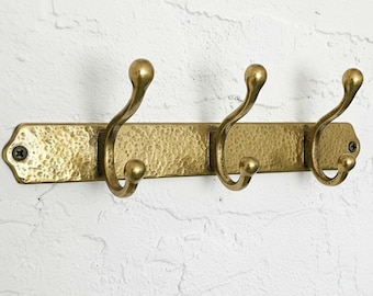 May include: A brass wall-mounted coat rack with three curved hooks. The rack has a hammered texture and is mounted on a white textured wall. The hooks are a classic design, suitable for a hallway or entryway.