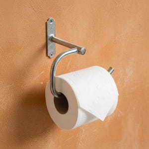 Puede incluir: Un portarrollos de papel higiénico de metal plateado montado en una pared texturizada de tono naranja. El soporte tiene un brazo curvo para sujetar el rollo de papel higiénico blanco. La base de montaje en pared es rectangular con dos tornillos.