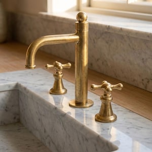May include: A gold-colored faucet with a curved spout and two cross-shaped handles. The faucet is mounted on a marble countertop, with a matching marble sink visible in the foreground. The design is classic and elegant.