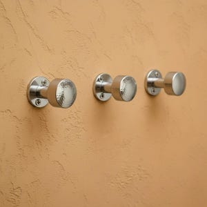 Könnte beinhalten: Drei silberfarbene Wandhaken mit gehammerter Textur auf der Vorderseite. Die Haken sind an einer beigefarbenen Wand montiert. Die ersten beiden Haken haben eine runde Form, während der dritte eine zylindrische Form hat.