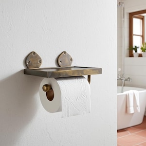 May include: A brass-coloured wall-mounted toilet roll holder with a small shelf. It holds a roll of white toilet paper. The design includes two decorative, teardrop-shaped mounting plates. The holder is designed for a bathroom.