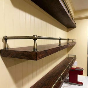 Puede incluir: Tres estantes de madera marrón oscuro con barandillas y soportes de metal color latón están montados en una pared color crema. Los estantes son de diferentes longitudes y están instalados a diferentes alturas. Los estantes están vacíos.