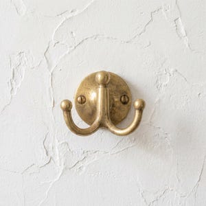May include: A brass double hook wall-mounted coat rack. The rack has a circular backplate with two screws and two curved hooks with rounded ends. The finish is a warm, antique brass color, and it is mounted on a textured white wall.