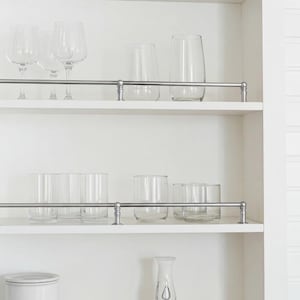 Op de afbeelding: Een witte kast met meerdere planken met diverse heldere drinkglazen. Wijnglazen, bekers en lowball-glazen zijn op de planken geplaatst. Een metalen stang loopt langs de voorkant van elke plank.