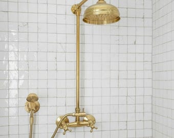 May include: A gold-colored shower fixture with a round showerhead and a handheld showerhead. The showerhead is mounted on a vertical pipe and has a curved arm. The fixture has two cross-shaped handles and is set against a white tiled wall.