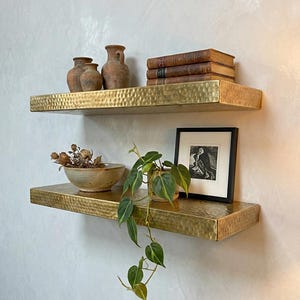 Puede incluir: Dos estantes flotantes de latón martillado montados en una pared gris claro. El estante superior contiene tres pequeños jarrones marrones y varios libros antiguos. El estante inferior exhibe un cuenco de cerámica, una impresión enmarcada y una planta en maceta.