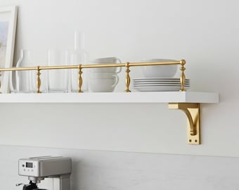 May include: A white floating shelf with a brass railing and brackets, holding white dishes, cups, and glasses. The shelf is mounted on a white wall above a stainless steel coffee maker.