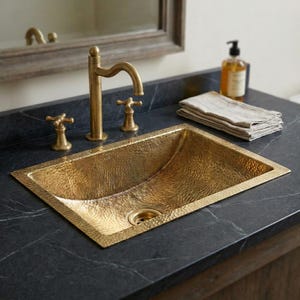 Op de afbeelding: Een rechthoekige gehamerde messing wastafel met bijpassende kraan en armaturen. De wastafel is ingebouwd in een donker aanrechtblad, met een stapel gevouwen handdoeken en een zeepdispenser in de buurt. De kraan en armaturen hebben een vintage messing afwerking.