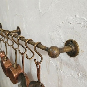 May include: Antique brass pot rack with S-hooks, displaying copper pans. The rack is fixed to a textured white wall. The design is classic, ideal for kitchen storage and organization.