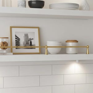 以下が含まれることがあります： 様々な装飾品が置かれた白いキッチン棚。 白黒写真が入った金色のフォトフレーム、クッキーが入ったガラス瓶、白いボウル、金色の棚レール。