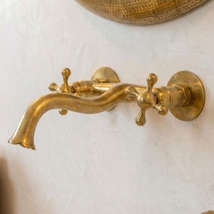 May include: A brass wall-mounted tap with a curved spout and cross-shaped handles. The tap has a vintage design and is mounted on a white wall. The fixture is made of brass and has a polished finish.