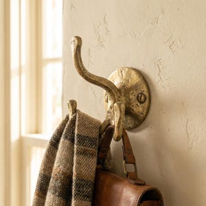May include: A brass wall hook with a double hook design. A brown and tan plaid scarf and a brown leather bag hang from the hooks. The hook is mounted on a textured, off-white wall.