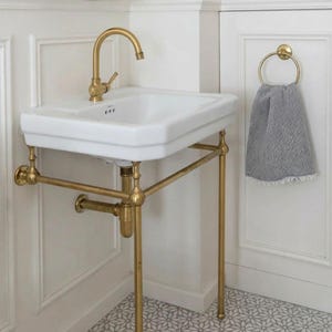 以下が含まれることがあります： 真鍮の蛇口と脚が付いた白い磁器の洗面台。真鍮のタオルリングには、グレーと白の模様のタオルがかかっています。洗面台は白いパネルの壁に設置され、模様入りのタイルの床に置かれています。