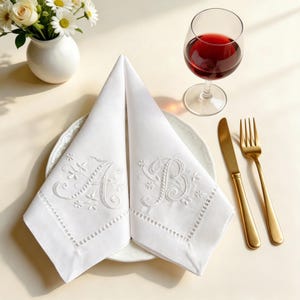 May include: Two white cloth napkins folded on a plate, each featuring white embroidered floral details and the letters A and B. The set is accompanied by a gold-toned knife and fork, a glass of red wine, and a small vase of white flowers.