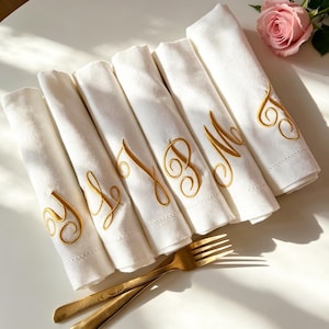 Può includere: Un set di sei tovaglioli in tessuto bianco, ognuno caratterizzato da una lettera corsiva ricamata in filo dorato. I tovaglioli sono disposti in fila, accompagnati da due forchette in metallo dorato. Una rosa rosa è posizionata nell'angolo in alto a destra.