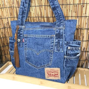 Può includere: Una borsa tote in denim blu realizzata con jeans riciclati, dotata di una tasca centrale con etichetta Levi Strauss & Co., tasche laterali e un ciondolo a nappa in pelle marrone con perline di legno su uno dei due manici.
