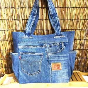 Puede incluir: Un bolso tote de mezclilla azul hecho de jeans reciclados, con dos correas para el hombro, bolsillos laterales y un bolsillo trasero con una etiqueta roja de Levi Strauss & Co. Un parche de marca color marrón está cosido en la parte inferior.