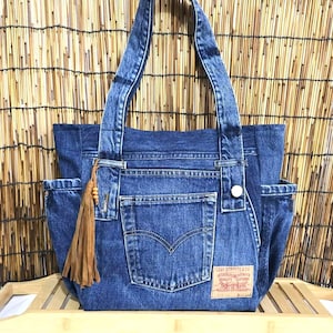 Può includere: Una borsa tote in denim blu con due tasche esterne e un nappa marrone. La borsa presenta un'etichetta Levi Strauss & Co. e una chiusura con bottone. La borsa ha due manici.