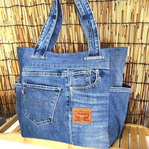 Może przedstawiać: Niebieska torba typu tote z dżinsu z dwoma uchwytami i wieloma kieszeniami. Torba posiada tylne kieszenie pary dżinsów, w tym etykietę Levi Strauss & Co. i czerwoną metkę. Torba jest wykonana z niebieskiego dżinsu.