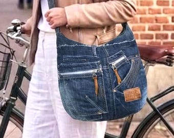 May include: A dark blue denim hobo bag featuring multiple exterior pockets with silver zippers, brown leather zipper pulls, a frayed top edge, and a rectangular brown leather patch on the lower right side, worn with a long adjustable strap.