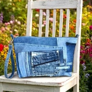 Pode incluir: Uma bolsa de mão retangular em ganga, construída com vários retalhos de tecido em tons de azul. A bolsa apresenta um bolso externo proeminente com costuras visíveis e uma alça de pulso em ganga a condizer, presa com um fecho de metal prateado.