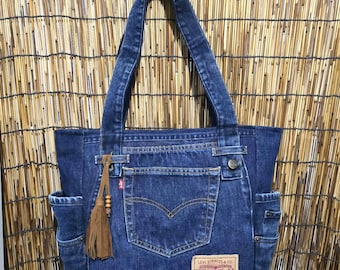 May include: A dark blue denim tote bag constructed from repurposed jeans, featuring a central back pocket, a Levi Strauss & Co. leather patch, two side pockets, and a brown suede tassel with wooden beads attached to one of the two shoulder straps.