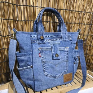 Op de afbeelding: Een blauwe denim tas gemaakt van gerecyclede jeans, voorzien van twee handvatten, een afneembare schouderriem, meerdere buitenzakken, een centraal vak met knoopsluiting, een klein rood label en een bruine leren merkpatch.