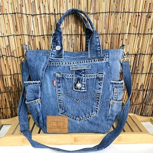 Könnte beinhalten: Eine blaue Tragetasche aus recyceltem Denim mit zwei Tragegriffen und einem abnehmbaren Schultergurt. Die Vorderseite zeigt eine mittige Tasche mit Knopfverschluss, ein kleines rotes Levi's-Markenetikett und einen beigen Lederaufnäher am Boden.